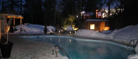 A swimming pool with a stone-tiled deck, surrounded by snow and trees.