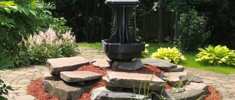 Un jardin agrémenté d’une fontaine en pierre, entourée de rochers et de paillis, avec une clôture en bois et des arbres en arrière-plan.