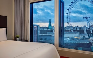 Une chambre d’hôtel dotée d’une grande fenêtre donnant sur un paysage urbain, avec notamment une tour imposante et une grande roue.
