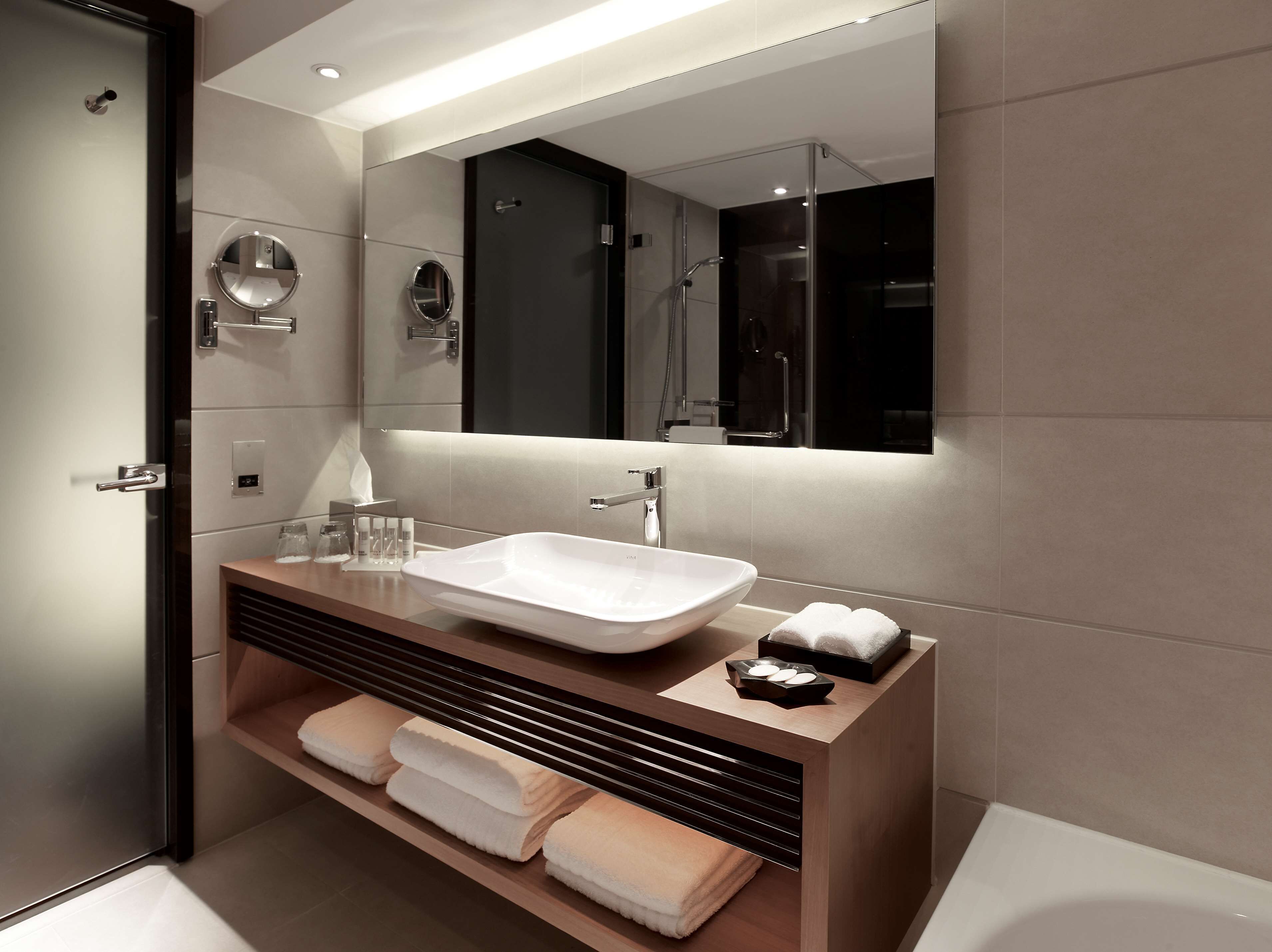 A modern bathroom with a large mirror, a white sink, and a wooden vanity.