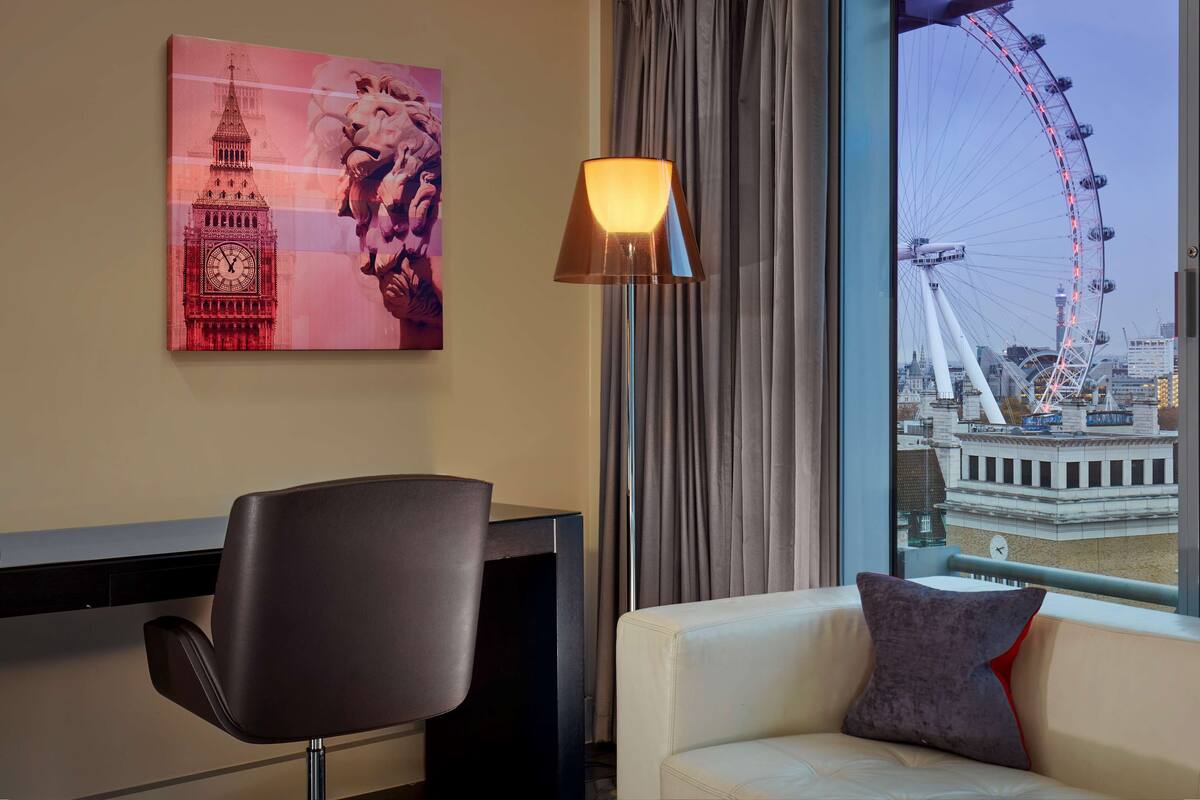 Una habitación de hotel con vistas a la London Eye, una pintura del Big Ben, una lámpara moderna y un sofá con un cojín.