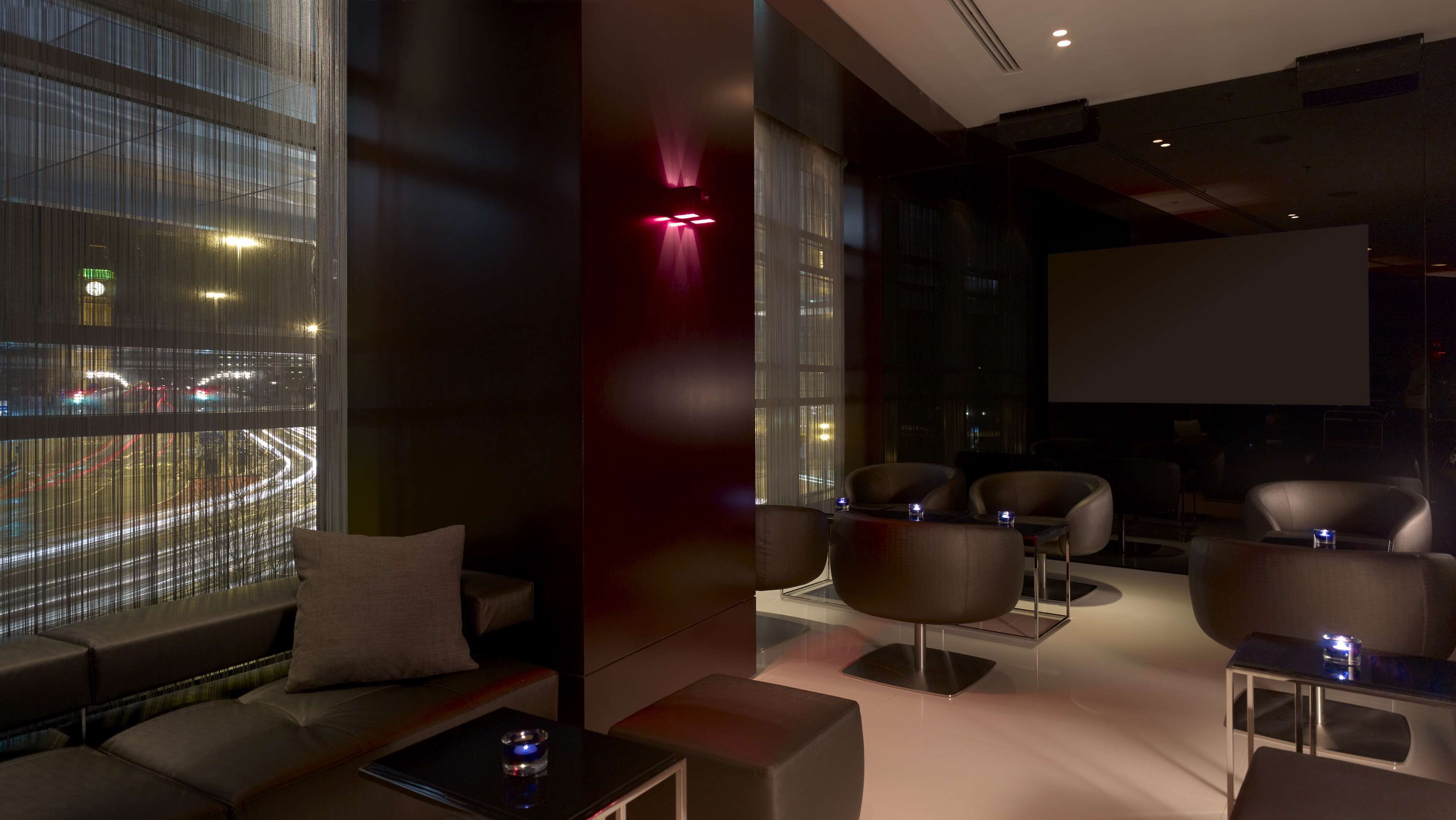 A modern lounge area with seating, a large window with city lights, and a projection screen.