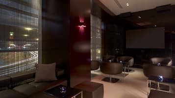 A modern lounge area with seating, a large window with city lights, and a projection screen.