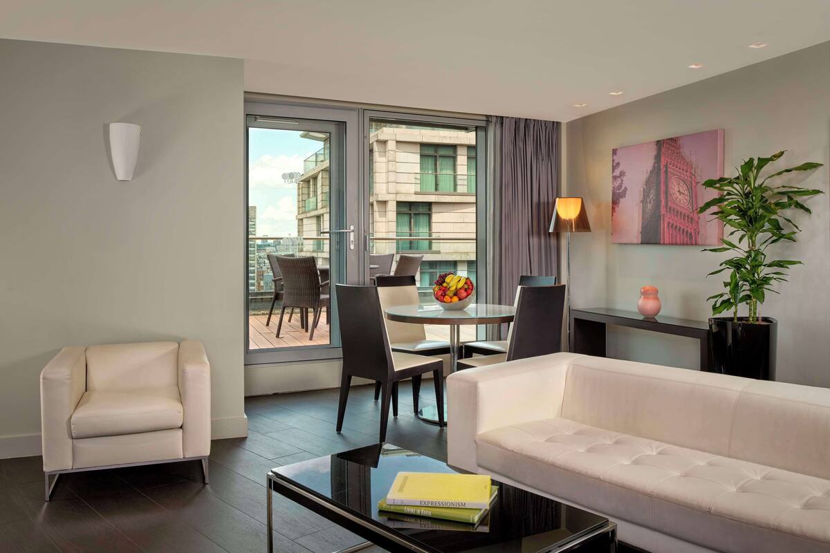 A modern living room with a sofa, armchair, dining table with chairs, a glass coffee table, a potted plant, and a painting of Big Ben.