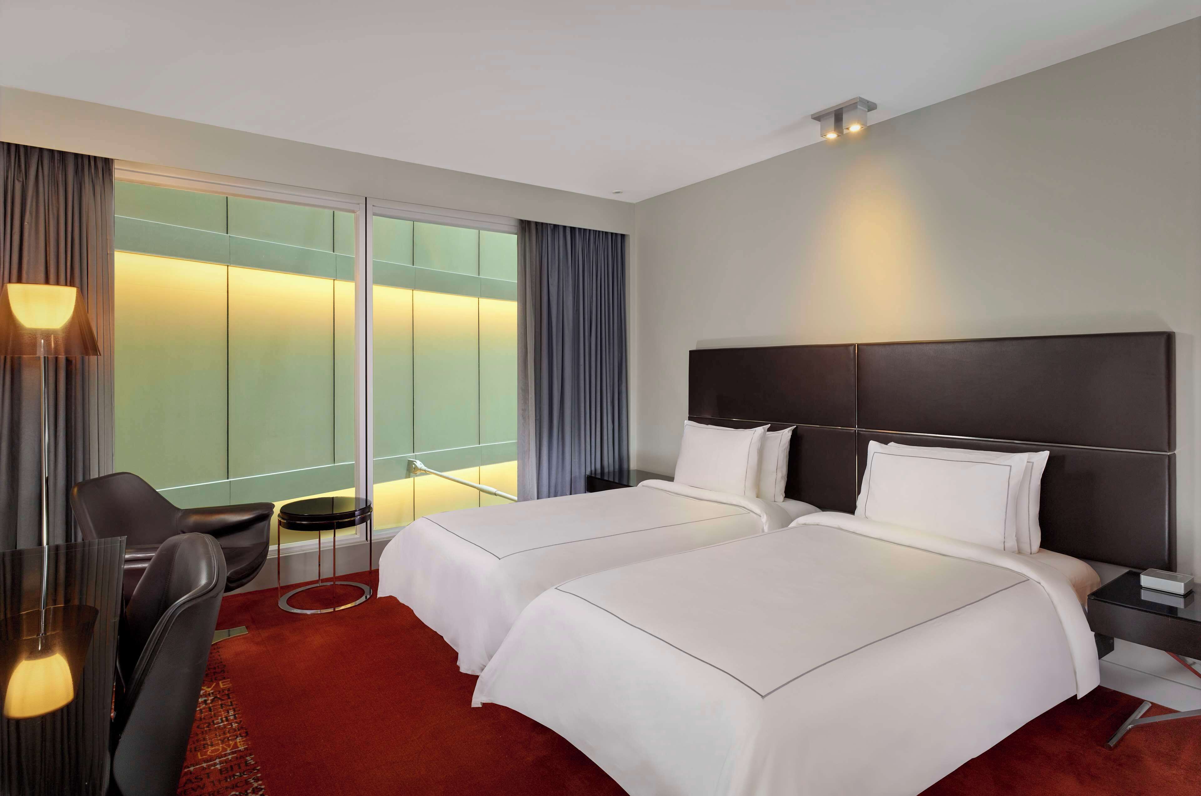 A hotel room with two beds, a desk, a chair, a small table, and a large window with curtains.