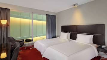 Quarto de hotel com duas camas, uma escrivaninha, uma cadeira, uma mesinha e uma janela grande com cortinas.