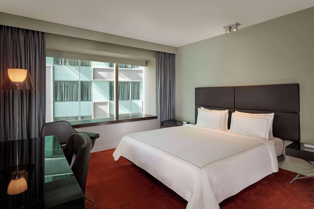 Una habitación de hotel con cama, escritorio, silla, ventana con cortinas y mesita de noche.