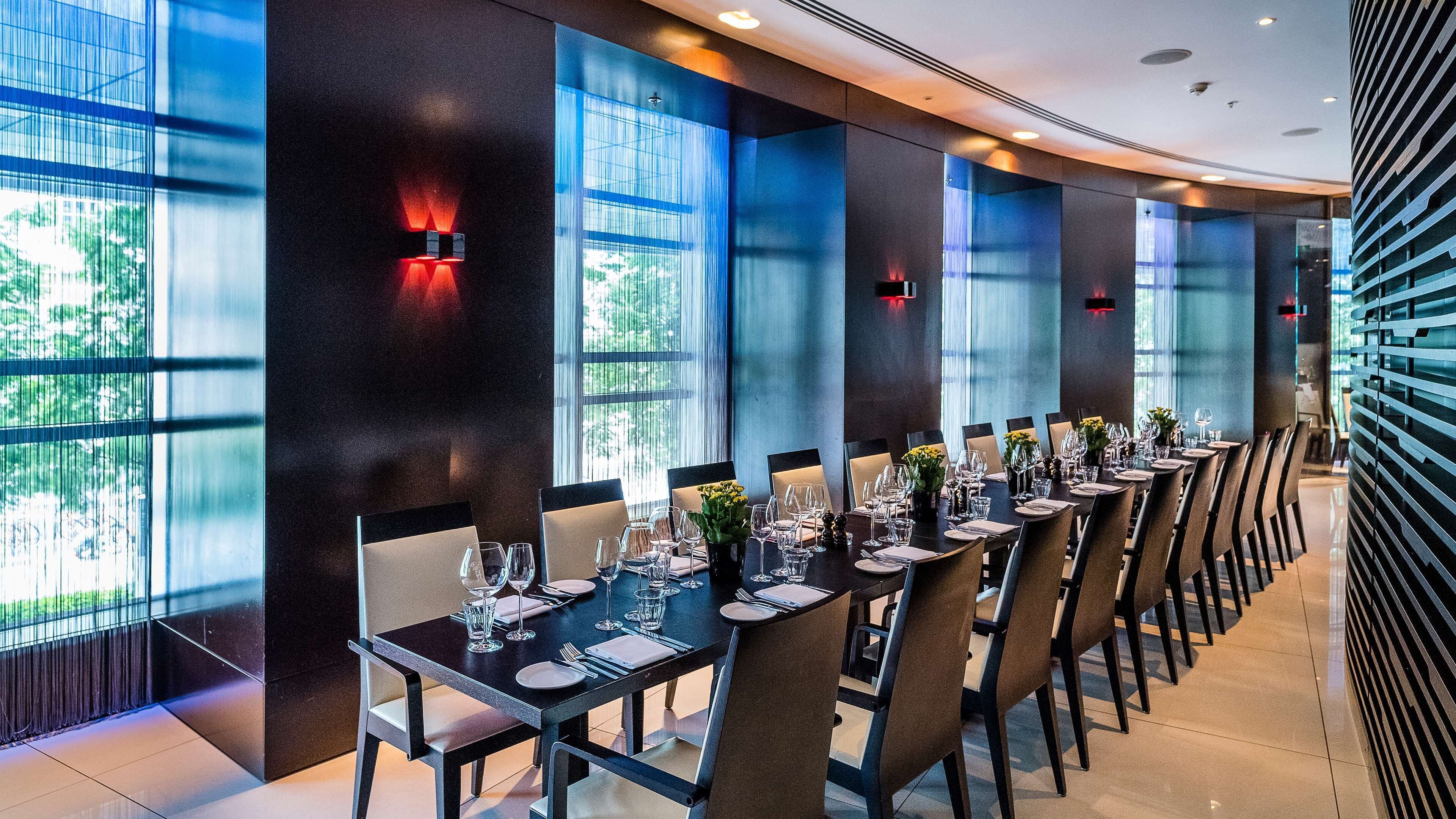 A modern dining area with long tables, chairs, and a view of greenery through large windows.