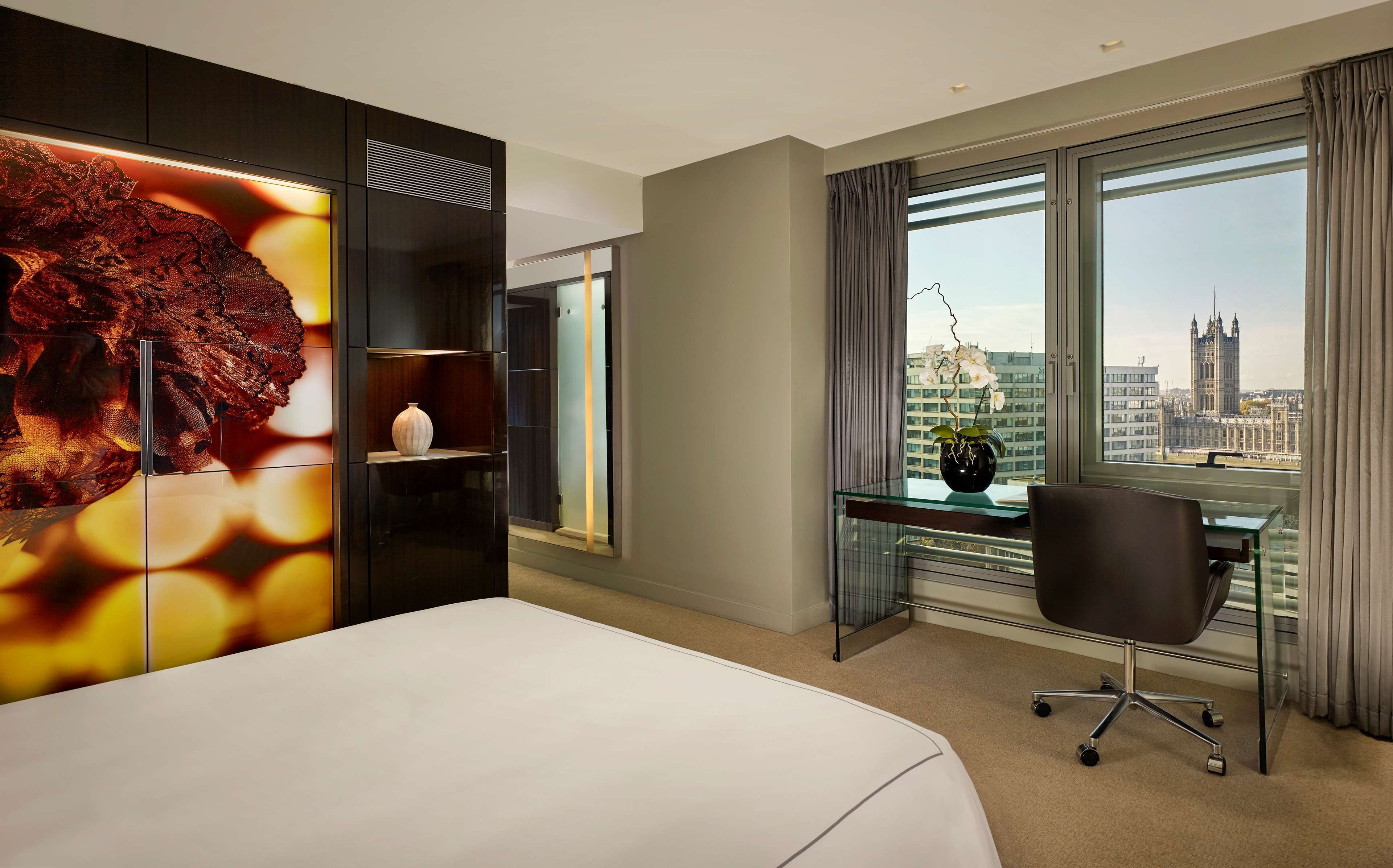 A hotel room with a bed, a desk, a chair, a large window with a city view, and a decorative wall piece.