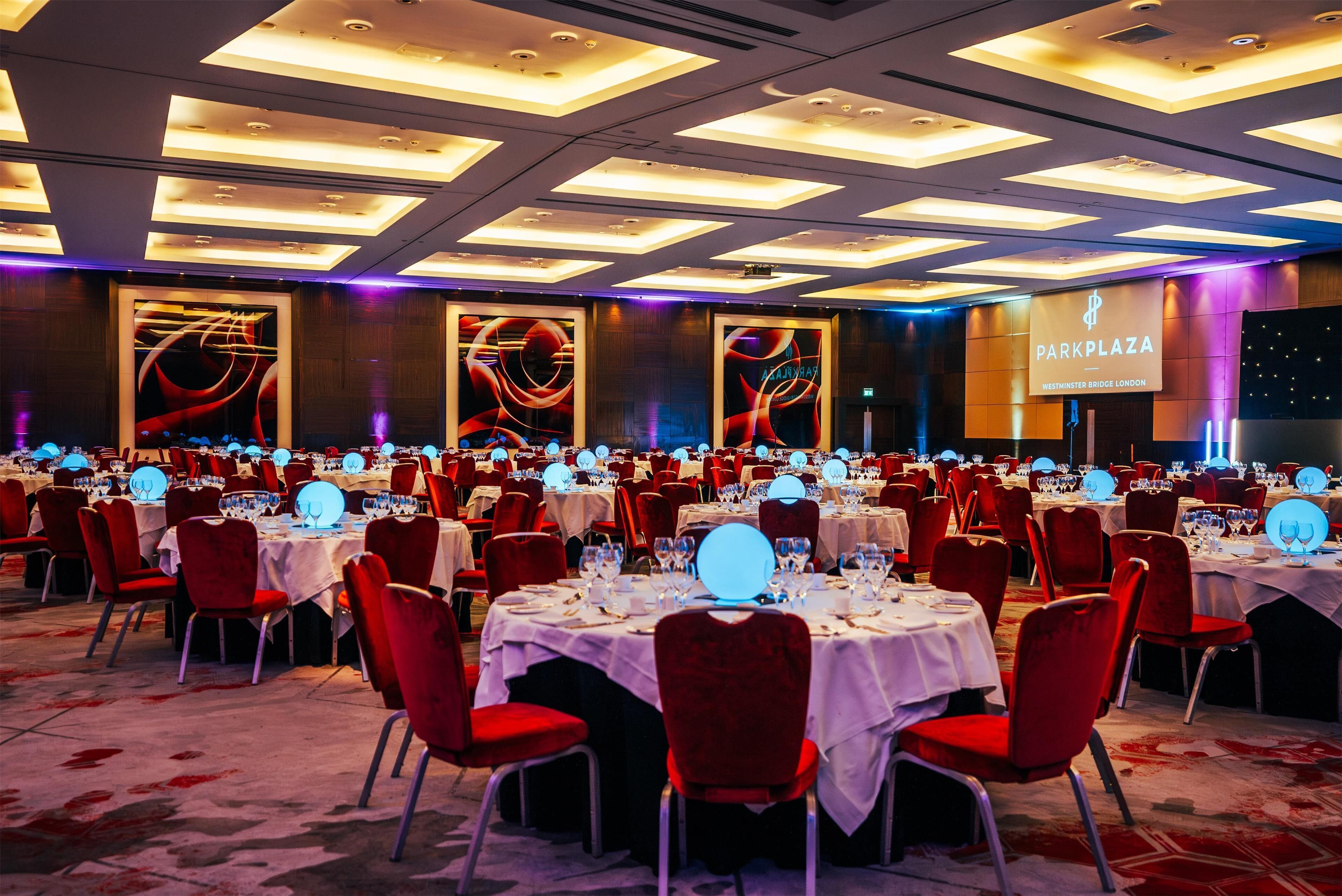 A banquet hall with round tables, red chairs, and a large screen displaying 'Park Plaza Westminster Bridge London'.