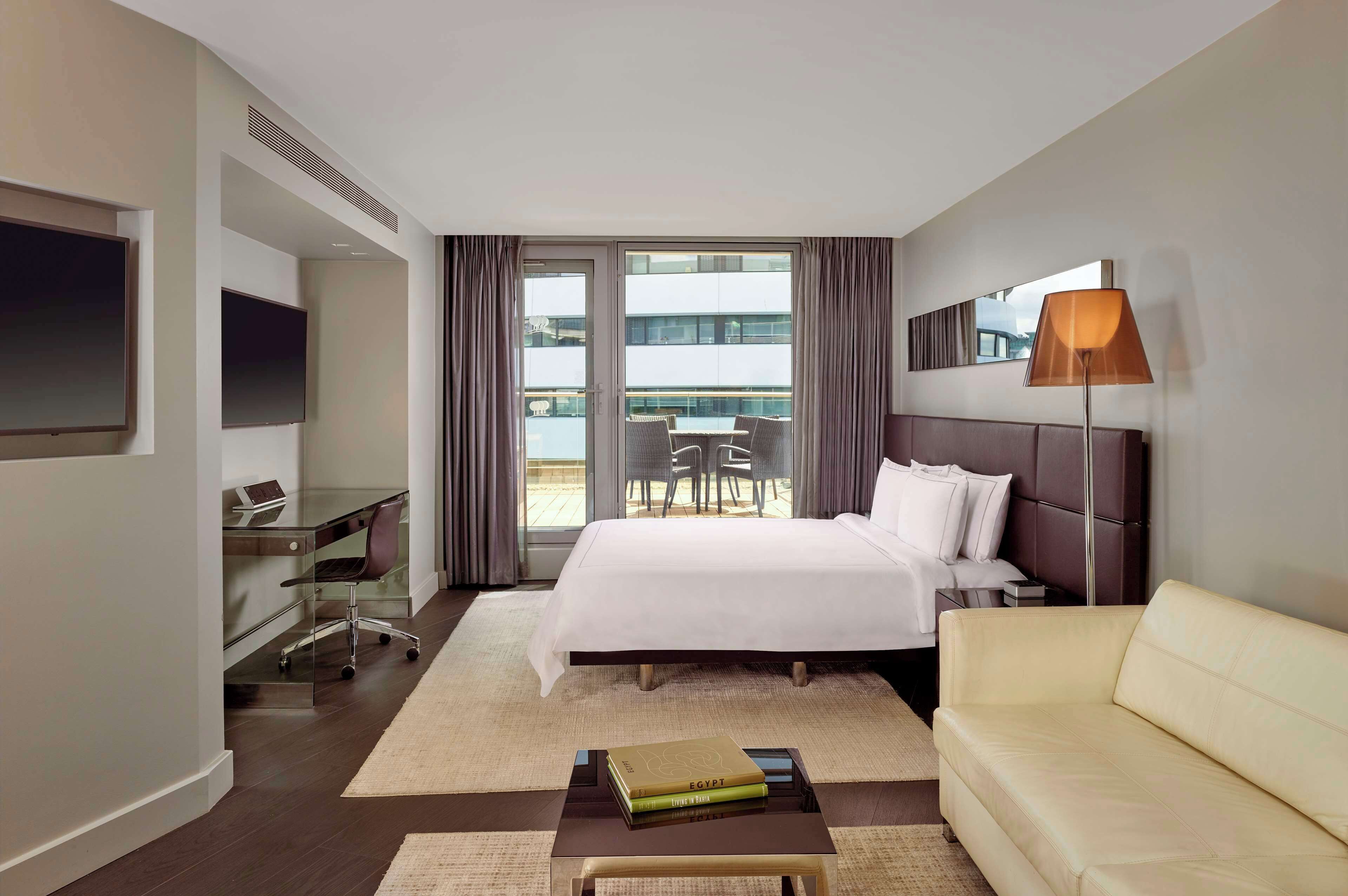 A hotel room with a bed, a desk, a TV, a sofa, and a balcony with a table and chairs.