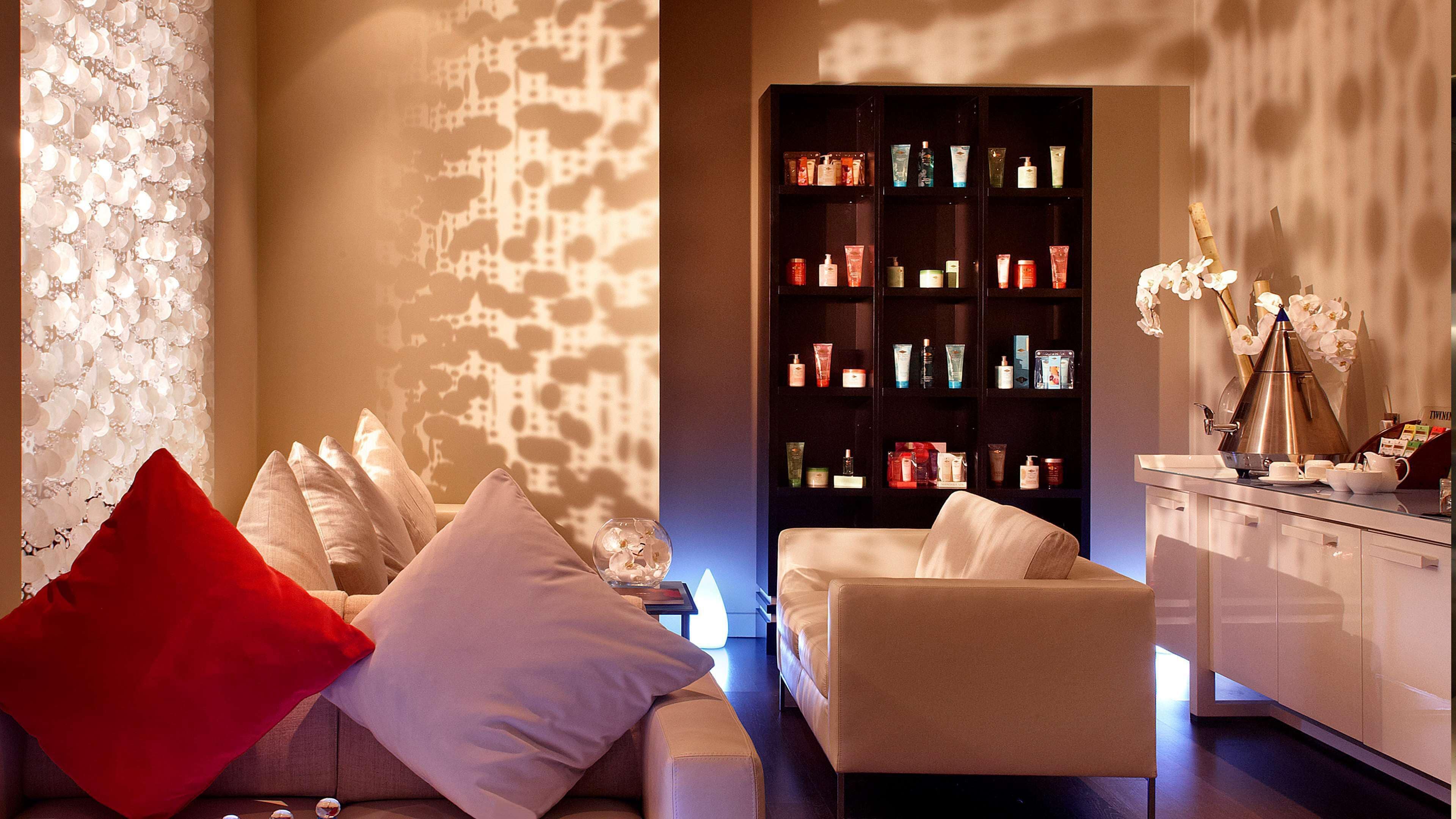 A modern room with a sofa, pillows, a bookshelf with toiletries, and a coffee maker.