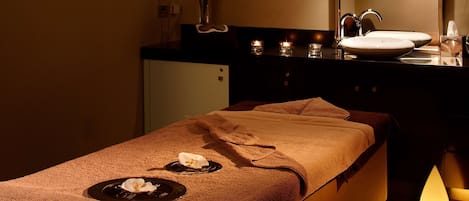 Ett massagebord med handdukar och en blomma, ett handfat med en spegel och ljus på en avställningsyta.