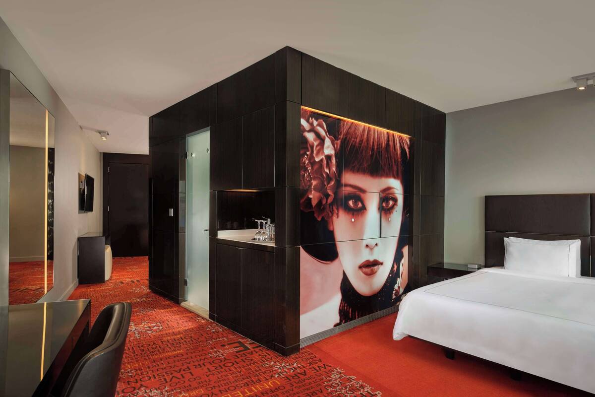 Una habitación de hotel moderna con una cama grande, un armario empotrado, una pequeña zona de estar y un mural grande en la pared.