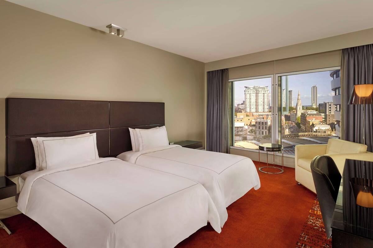Habitación de hotel con dos camas, un escritorio, una silla, un ventanal grande con vistas a la ciudad y una lámpara moderna.