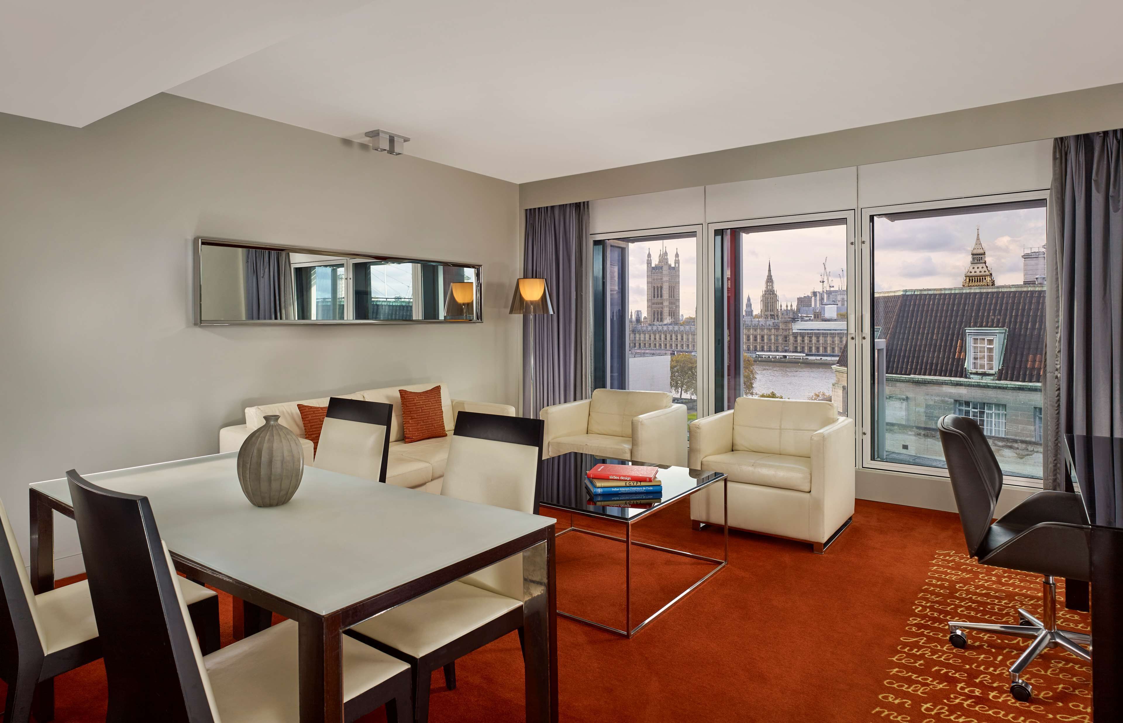 A modern hotel room with a dining area, a sofa, a coffee table, and a view of a cityscape.
