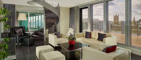 Ein modernes Wohnzimmer mit einer Sofagarnitur, einem Couchtisch, einer roten Vase mit Blumen, einer Treppe und Blick auf eine Stadtlandschaft durch große Fenster.