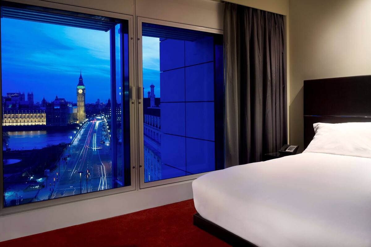 A hotel room with a large window offering a view of a cityscape, including a prominent clock tower, and a neatly made bed.