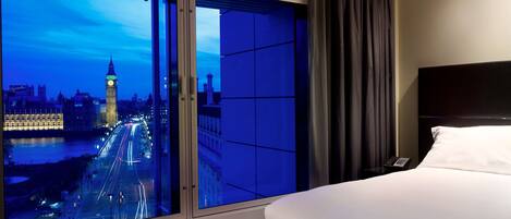 A hotel room with a large window offering a view of a cityscape, including a prominent clock tower, and a neatly made bed.