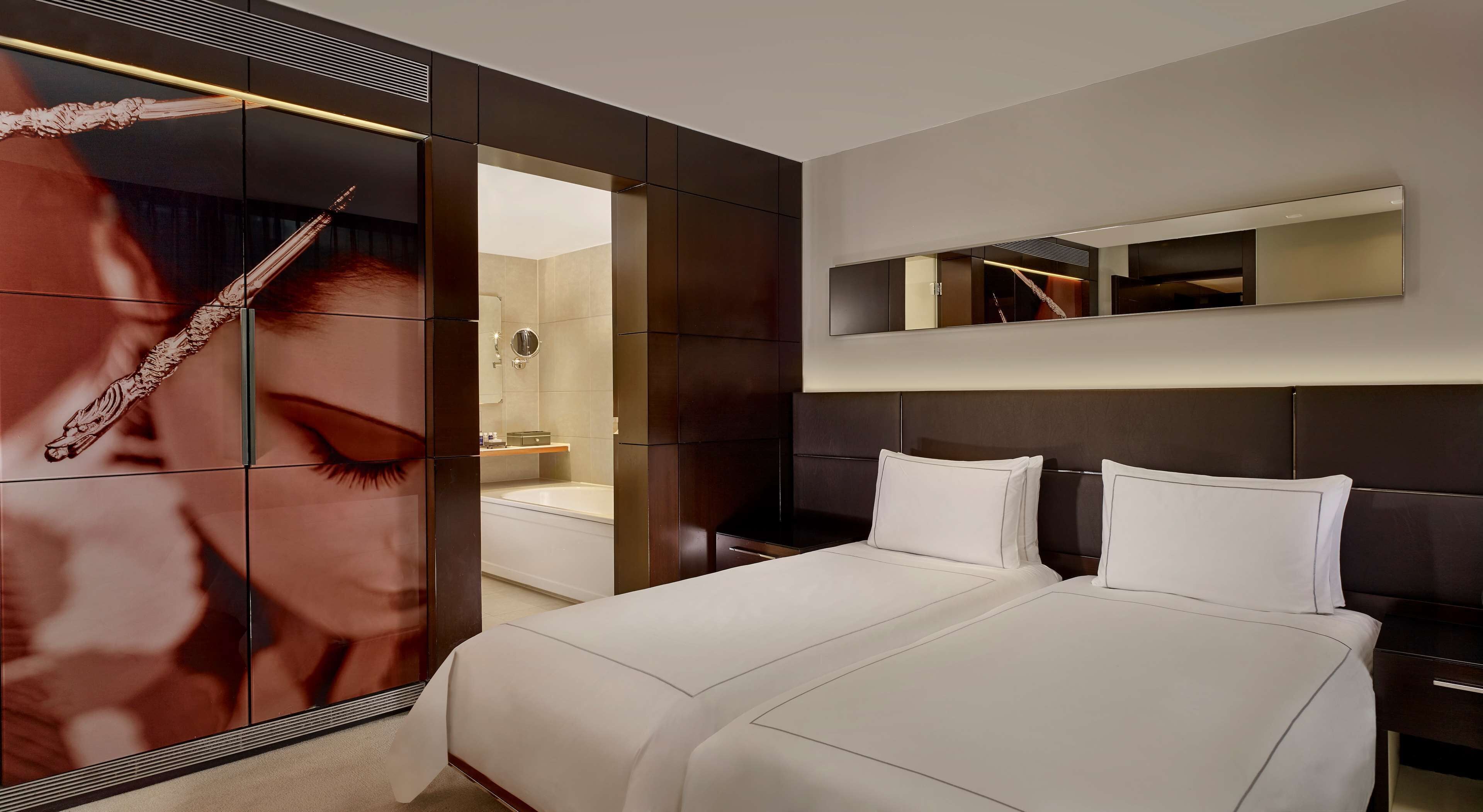 A modern hotel room with a large bed, a mirror above the bed, and a bathroom visible through a glass partition.