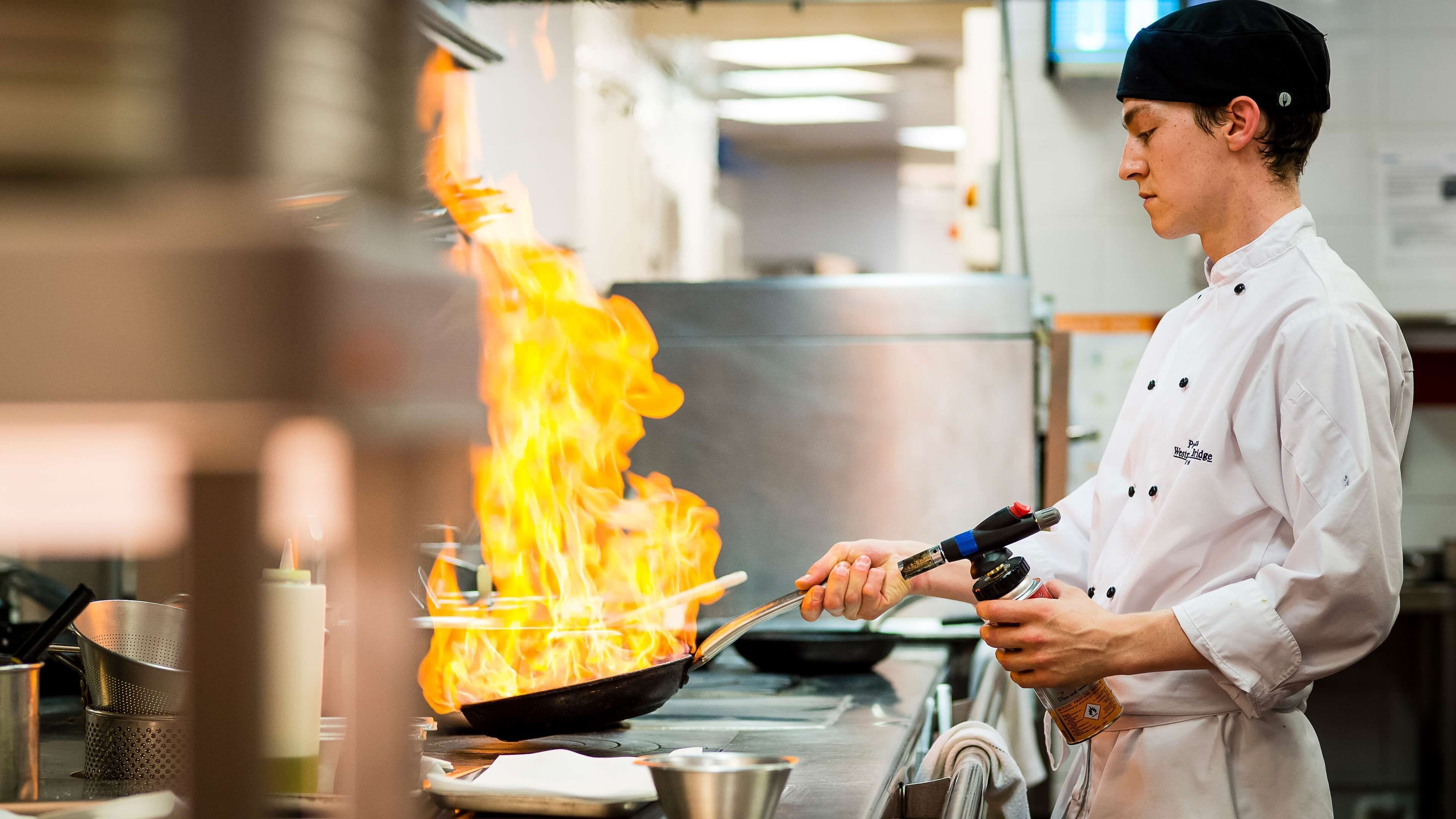 A chef in a professional kitchen with a pan on fire, using a fire extinguisher.