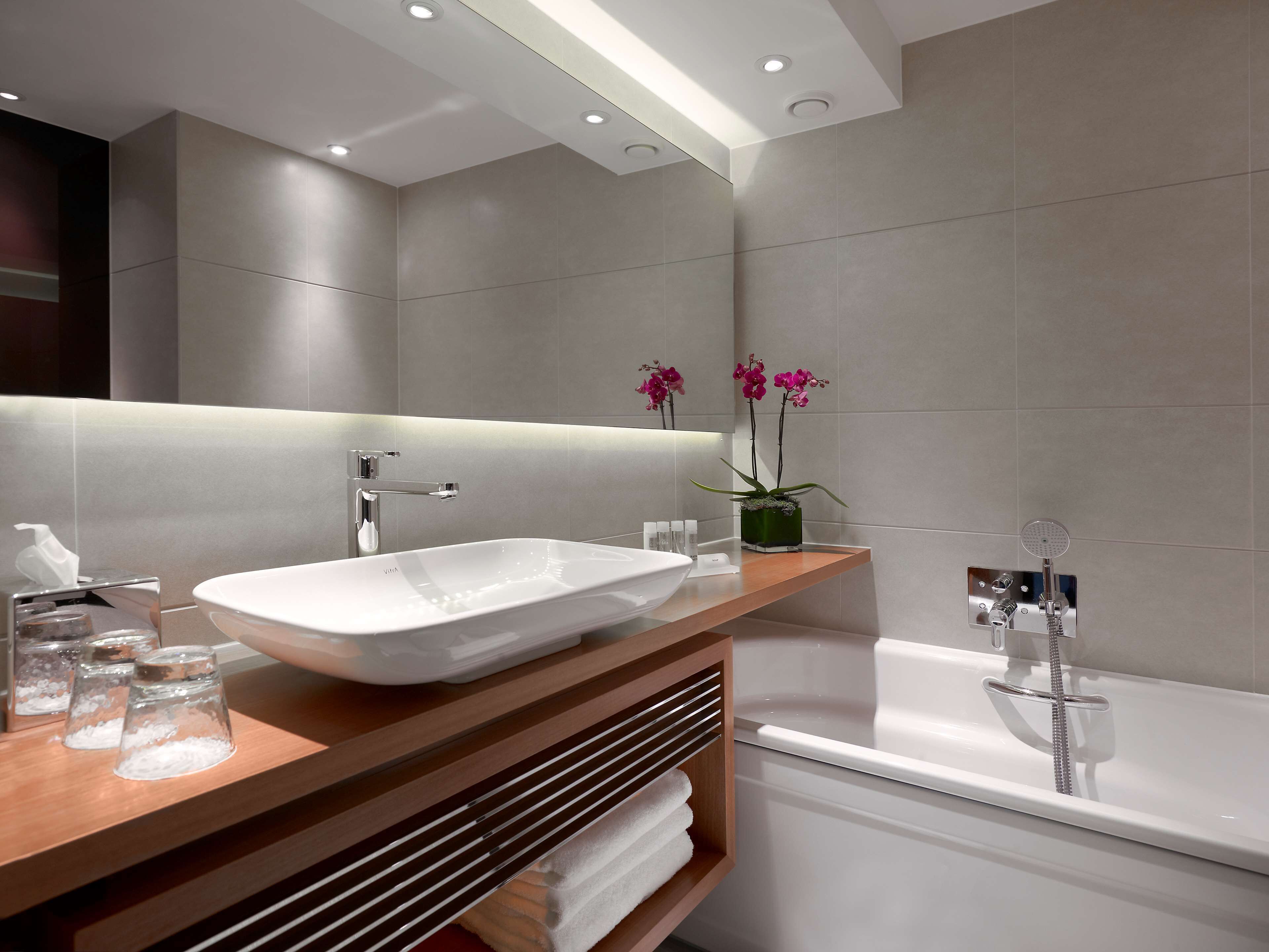 A modern bathroom with a large white sink, a wooden vanity, a built-in mirror, a shower with a handheld showerhead, and a potted orchid plant.