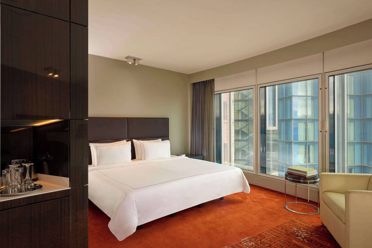 Una habitación de hotel con una cama grande, una zona de estar con una silla, una mesita pequeña y un ventanal con vistas a la ciudad.