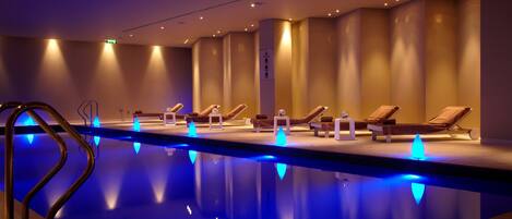 A swimming pool with blue lights, lounge chairs, and a tiled floor.