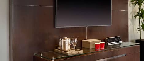 A modern reception desk with a glass top, a flat-screen TV mounted on the wall, a coffee pot, wine glasses, and a laptop.