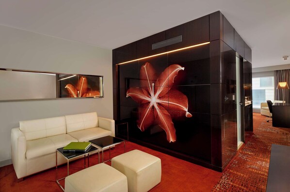 A modern hotel room with a white leather sofa, a glass partition with a large floral artwork, a red carpet, and a view of the city.