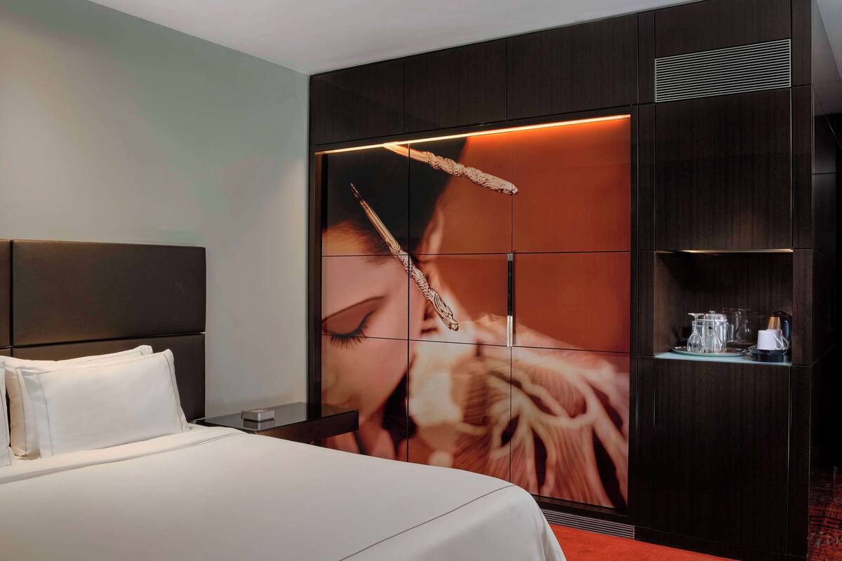 Una habitación de hotel con una cama grande, una mesita de noche, un estante en la pared con vasos y una cafetera, y un panel decorativo.
