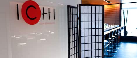 Un interior moderno con una barra de sushi y sashimi, una pared blanca con un logotipo rojo y un tradicional panel japonés.