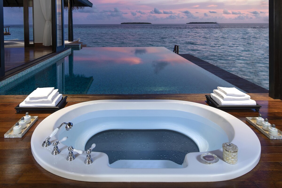 Una piscina infinita de lujo con vista al océano y al atardecer, un área de spa con una bañera grande, y toallas y artículos de aseo ordenados.