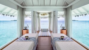 A room with two beds, a wooden floor, a large window with curtains, a hanging light fixture, and a view of the ocean. - Anantara Kihavah Maldives Villas (Kihavah)