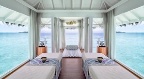 Una habitación con dos camas, suelo de madera, un ventanal grande con cortinas, una lámpara colgante y vista al océano.