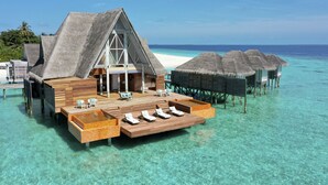 An overwater bungalow with a wooden deck, lounge chairs, and thatched-roof structures. - Anantara Kihavah Maldives Villas (Kihavah)
