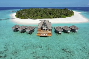 Cabañas sobre el agua con una terraza de madera central, rodeadas por aguas turquesas cristalinas y una playa de arena blanca.