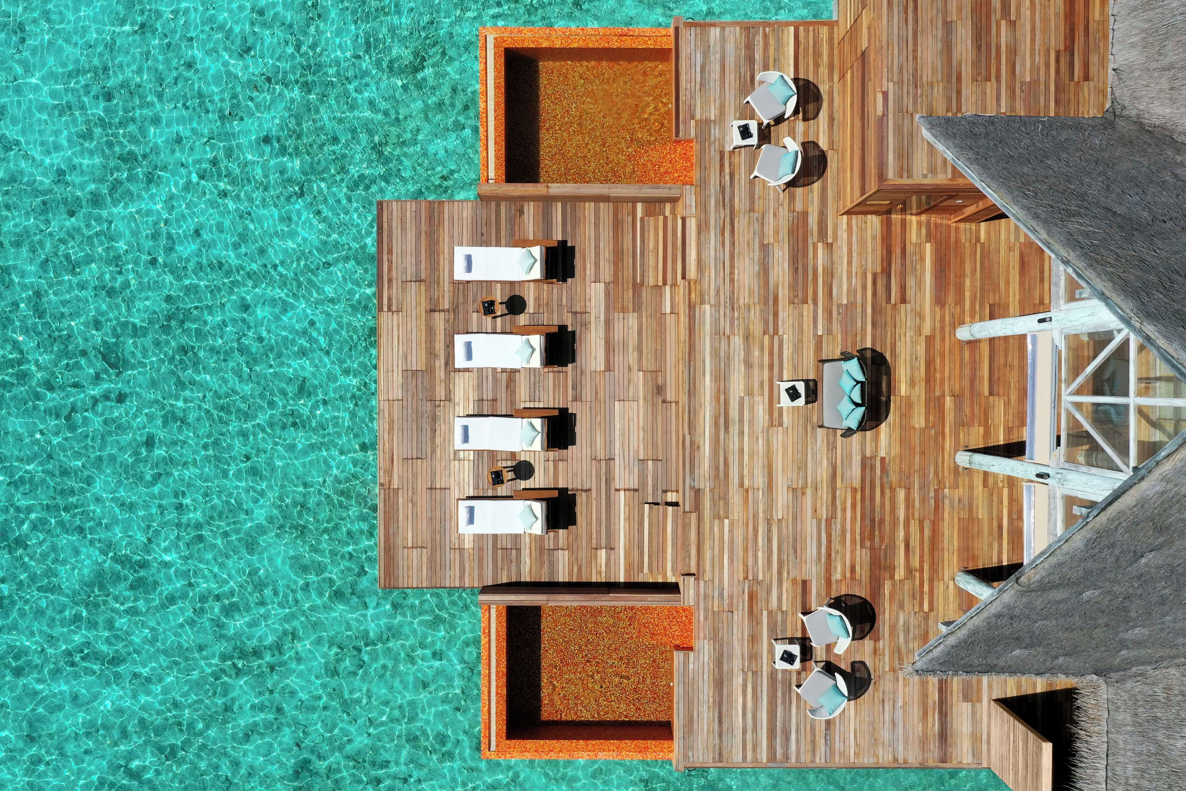 An aerial view of a wooden deck with lounge chairs and a small table, surrounded by clear turquoise water.