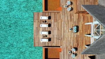 An aerial view of a wooden deck with lounge chairs and a small table, surrounded by clear turquoise water.