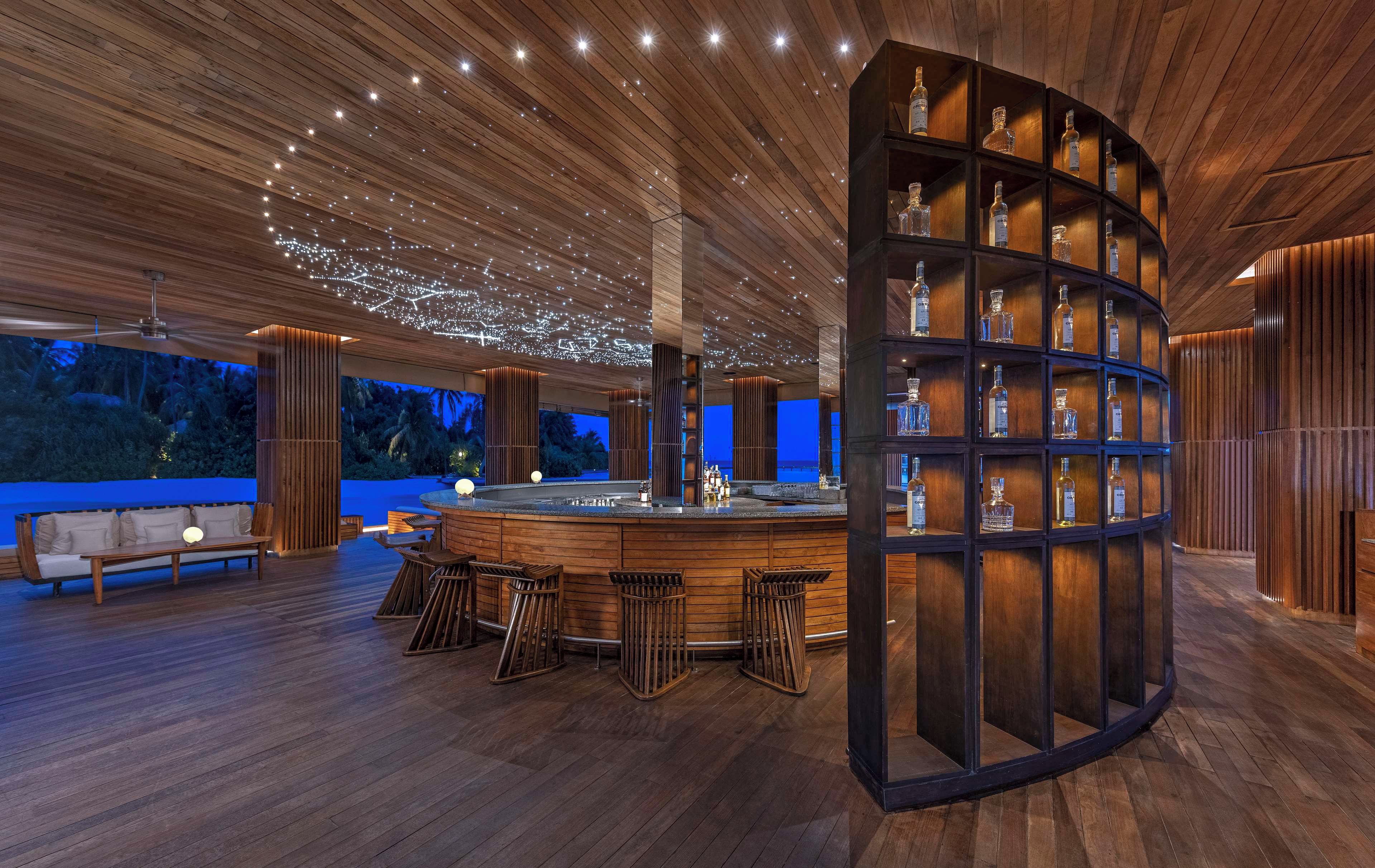 Zona de bar moderna con techo de madera, taburetes y estante con botellas.
