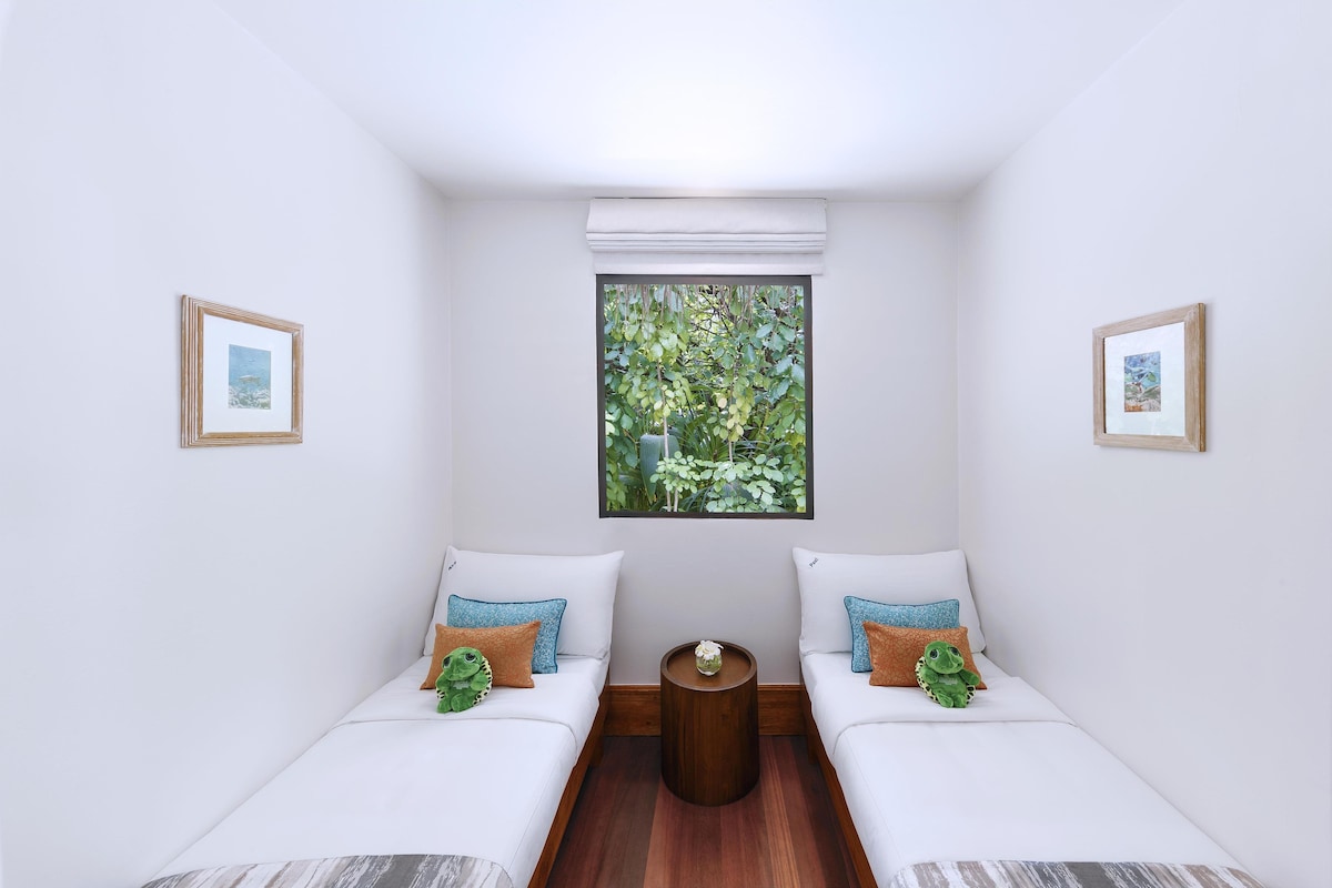 Una habitación pequeña y luminosa con dos camas blancas, una mesa redonda de madera y un ventanal con vistas a la vegetación.