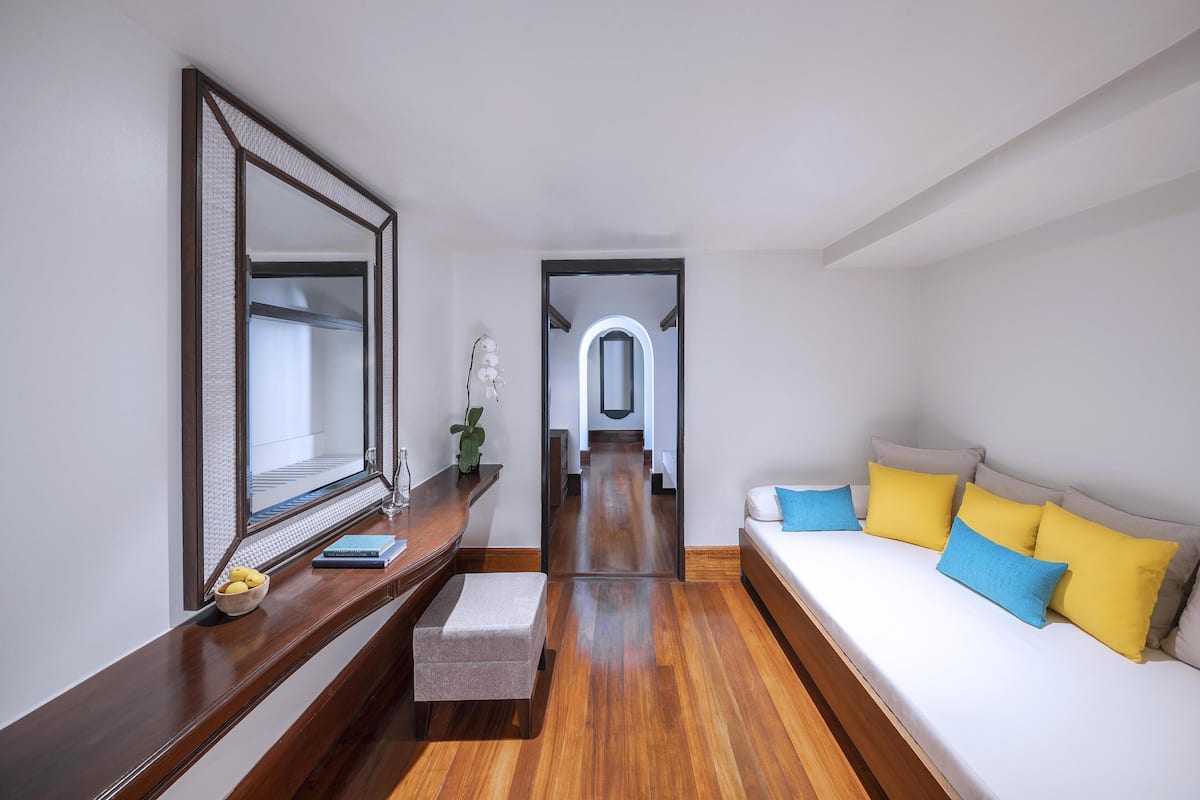 A modern hotel room with a wooden floor, a large mirror, a sofa with yellow and blue cushions, and a small table with a bowl of fruit.