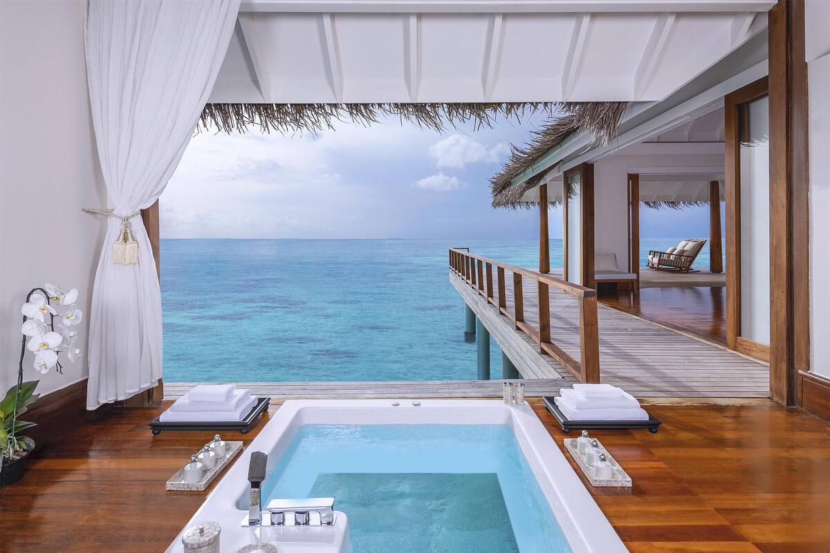 A luxurious room with a private balcony overlooking the ocean, a spa bathtub, and a wooden deck with lounge chairs.