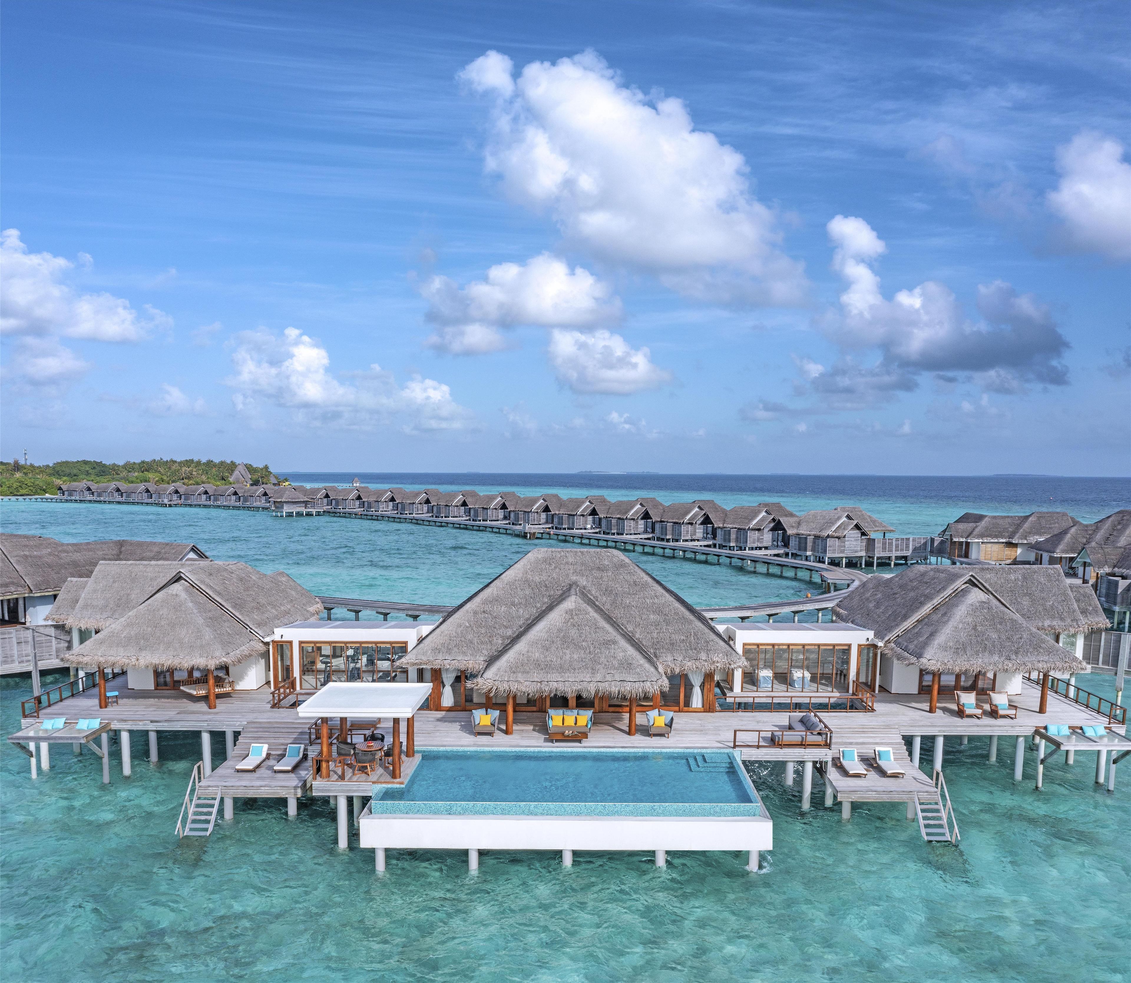 Des bungalows sur pilotis avec toits de chaume, une piscine centrale et une eau turquoise cristalline.
