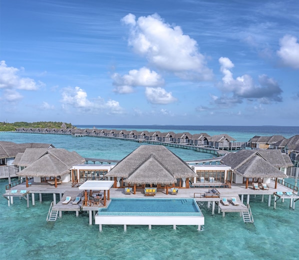 Overwater bungalows with thatched roofs, a central pool, and clear turquoise waters.
