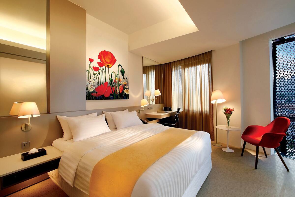 Una habitación de hotel con una cama grande, lámparas de noche, un escritorio, una silla, un cuadro y una ventana con cortinas.