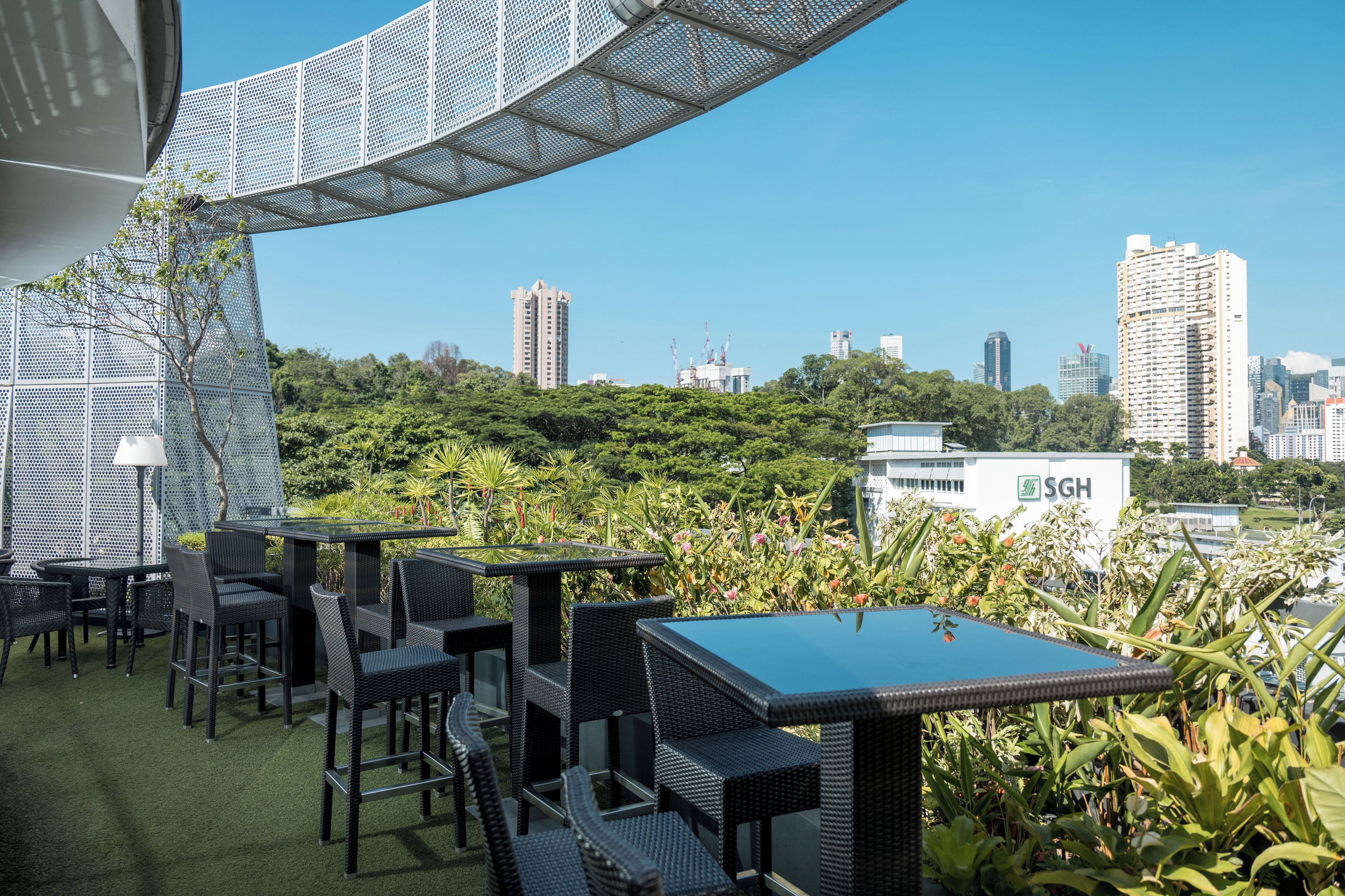 An outdoor seating area with tables and chairs, surrounded by greenery and overlooking a cityscape with high-rise buildings.