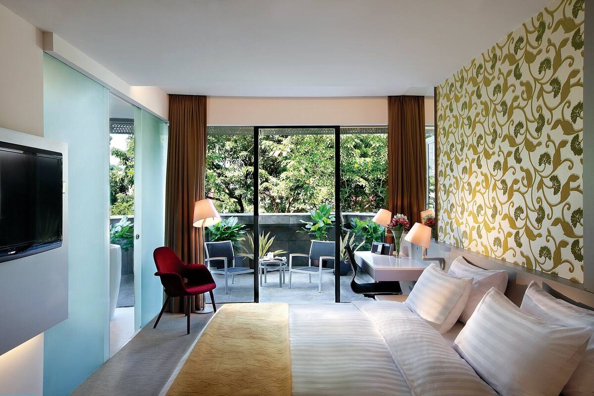 Habitación de hotel con cama, televisión, balcón con mesa y sillas, y vista a zonas verdes.