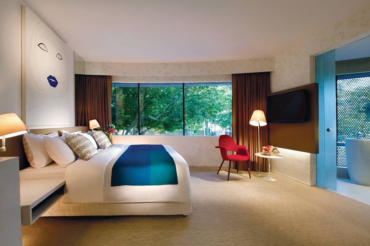 Habitación de hotel con una cama grande, un televisor montado en la pared, una mesita de noche con lámpara y una ventana con cortinas.
