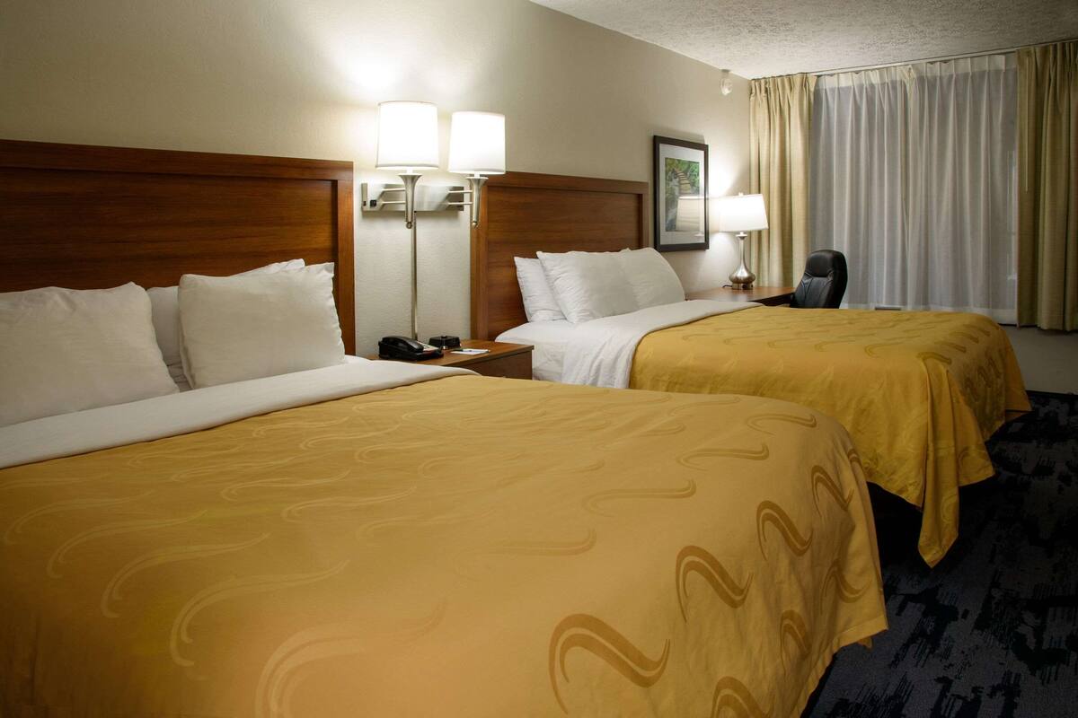 A hotel room with two beds, each with white linens and a yellow patterned bedspread. There are bedside lamps, a nightstand with a phone, and a chair.